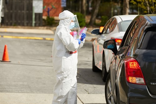 A person in white hazmat suit, face mask and face shield hands a ziplock bag to a driver in a black car.