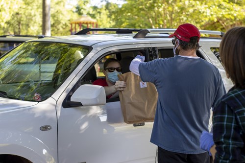 Man in red cap and blue t-shirt passes a paper bag through a car window to a driver wearing a face mask