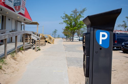 A parking meter in the foreground with a boardwalk along a beach and on-street parking.