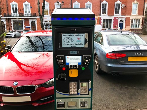 Parking meter in between a red and a black parked car, redbrick houses behind