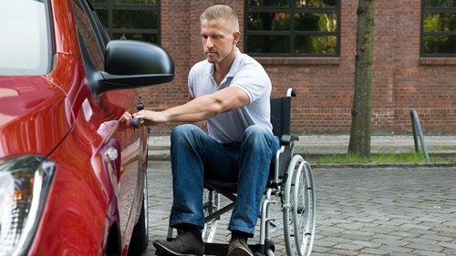 A man wearing pale polo shirt and jeans in a wheelchair opens the door of a red car
