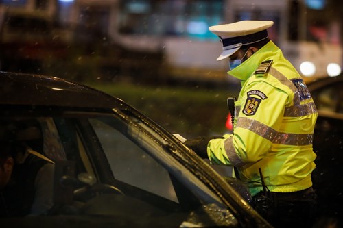 A parking enforcement officer wearing high-visibility jacket and surgical face mask issues a ticket to a black car.