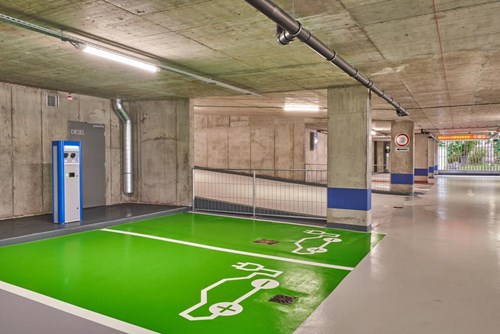 Parking spaces with green floor show electric vehicle icon and EV charging unit stands against the parking garage wall.