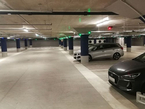 Inside a parking garage - a silver and black car parked in bays with green and red lights on the ceiling to show free/occupied spaces