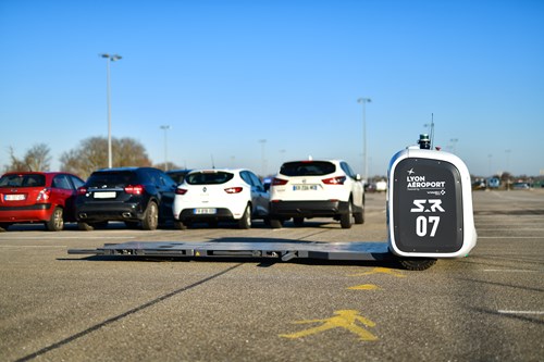A parking robot with flatbed to lift a car is parked in a car park with a row of four cars behind and a pedestrian painted on the tarmac in front